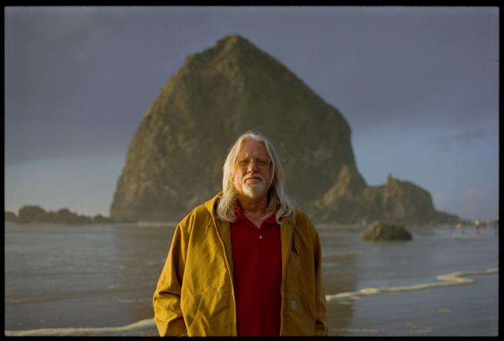 A man with long white hair and a beard stands on a beach with a large rock formation in the background.