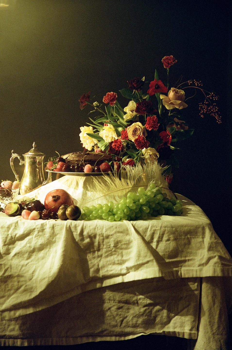 A still life arrangement features a bouquet of flowers, a cake, fruit, and a silver pitcher on a draped table.