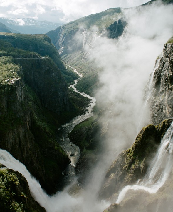 A river winds through a deep, green valley with waterfalls cascading down the misty cliffs.