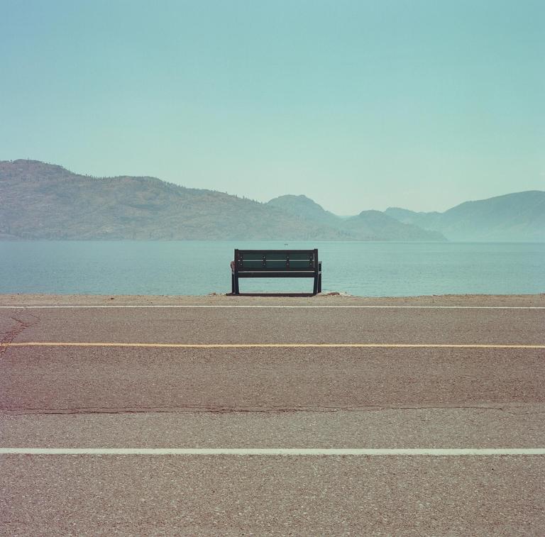 A lone bench sits beside a road overlooking a large body of water with mountains in the distance.