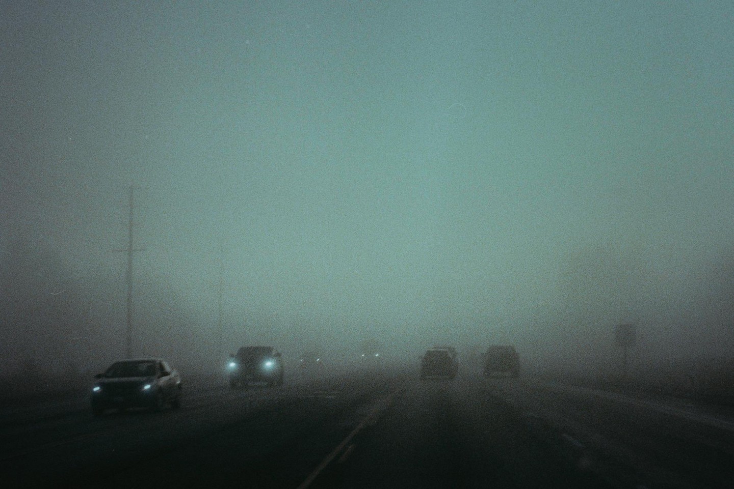 Cars drive on a road shrouded in thick fog, with their headlights cutting through the haze.