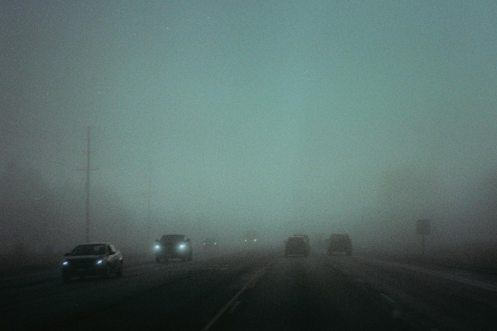 Cars drive on a road shrouded in thick fog, with their headlights cutting through the haze.