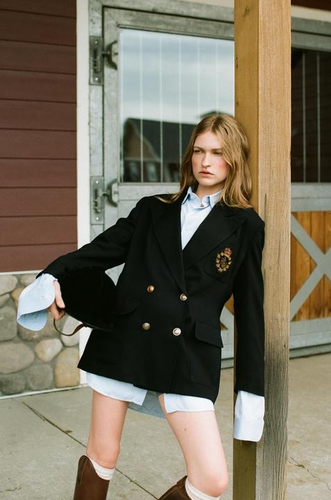 A woman in a blazer and boots leans against a wooden post next to a barn door.