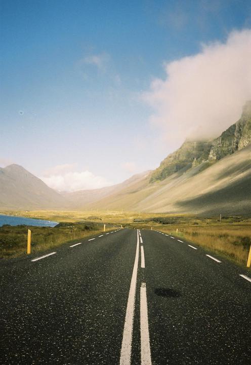 A long road stretches into the distance, flanked by mountains and a body of water under a blue sky.