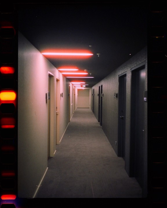 A long hotel hallway is illuminated by red neon lights on the ceiling.