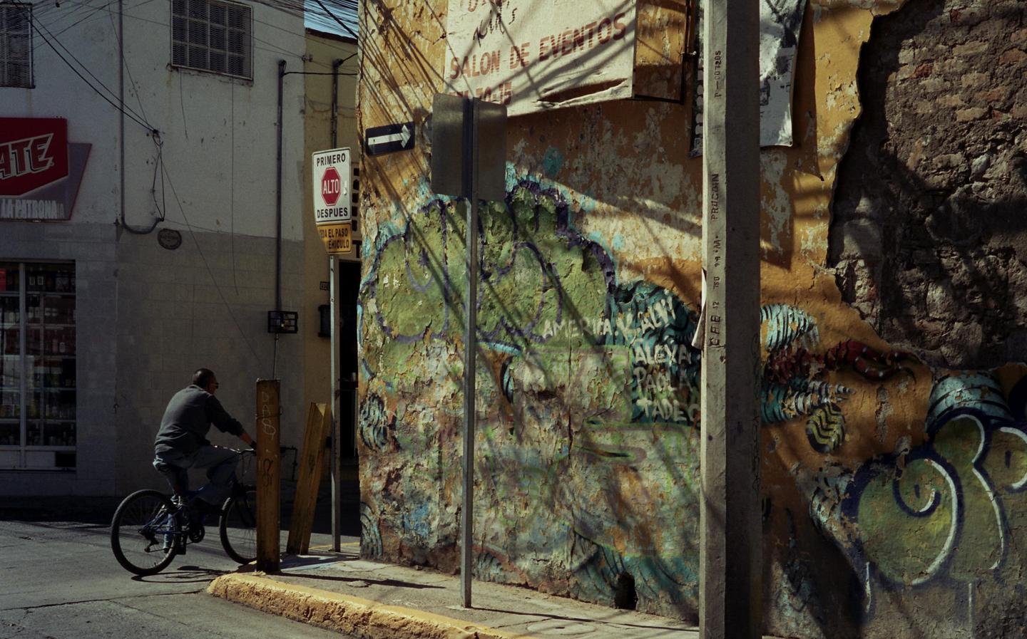 A man rides a bicycle past a building with peeling paint and graffiti on a sunny street.