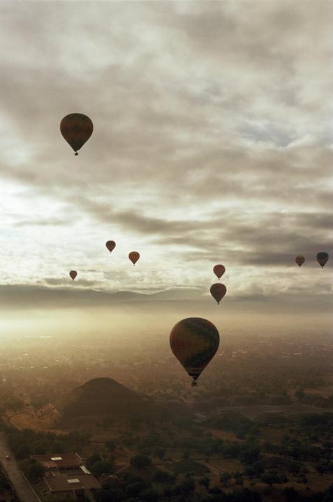 Hot air balloons float above a hazy landscape with a pyramid and mountains under a cloudy sky.