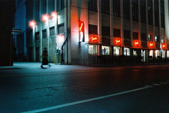 A blurry figure walks past a building with multiple neon signs that say "Fran's" at night.