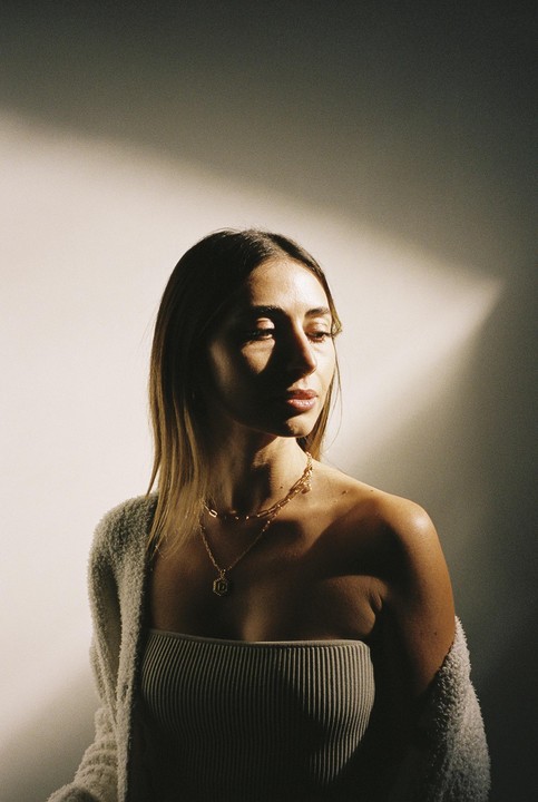 A woman with long hair and a ribbed top looks down, with a shadow cast across her face and the wall behind her.