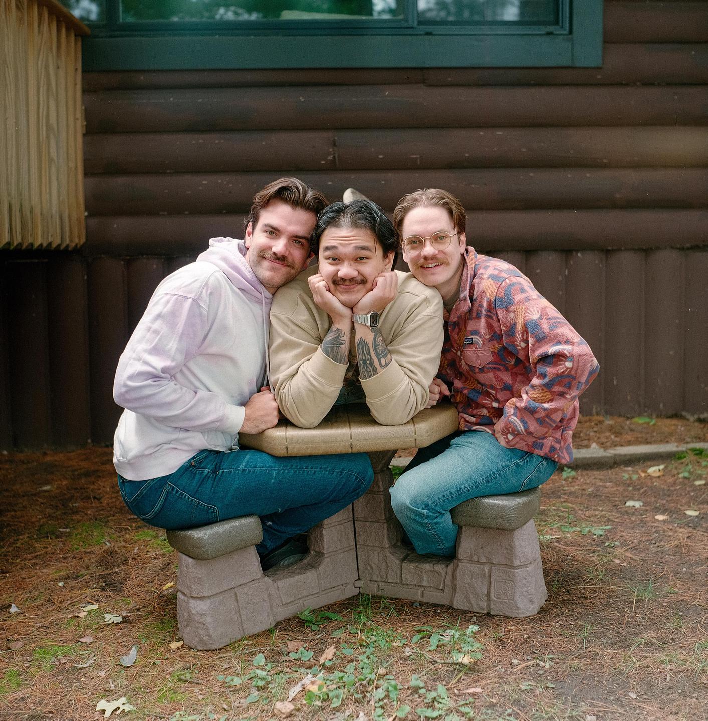Three men with mustaches are sitting at a small picnic table in front of a wooden cabin.
