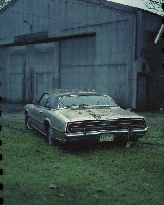 A dirty, old car is parked on a grassy area in front of a large, grey building.
