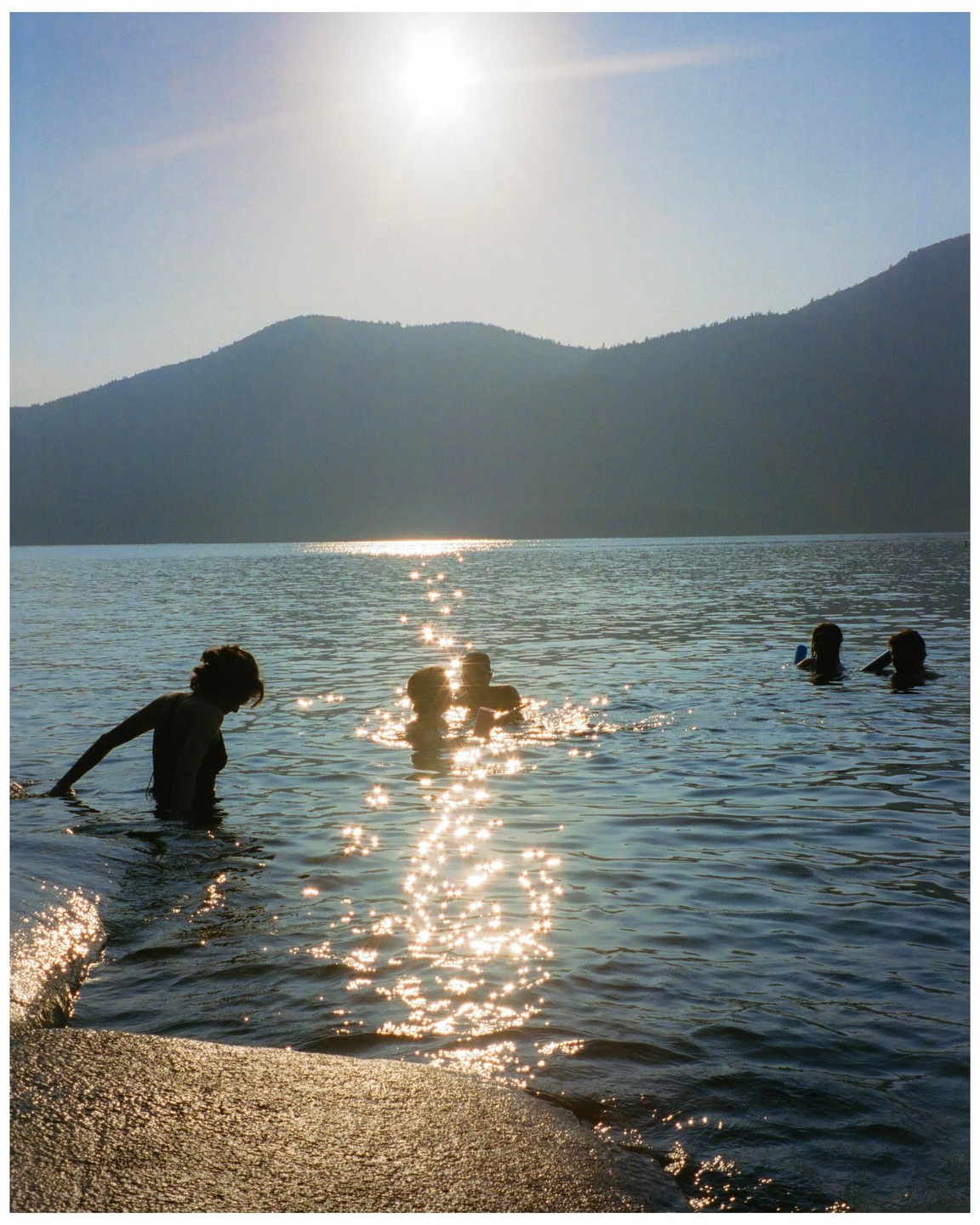 People swim in a lake with mountains in the background and sunlight reflecting on the water.