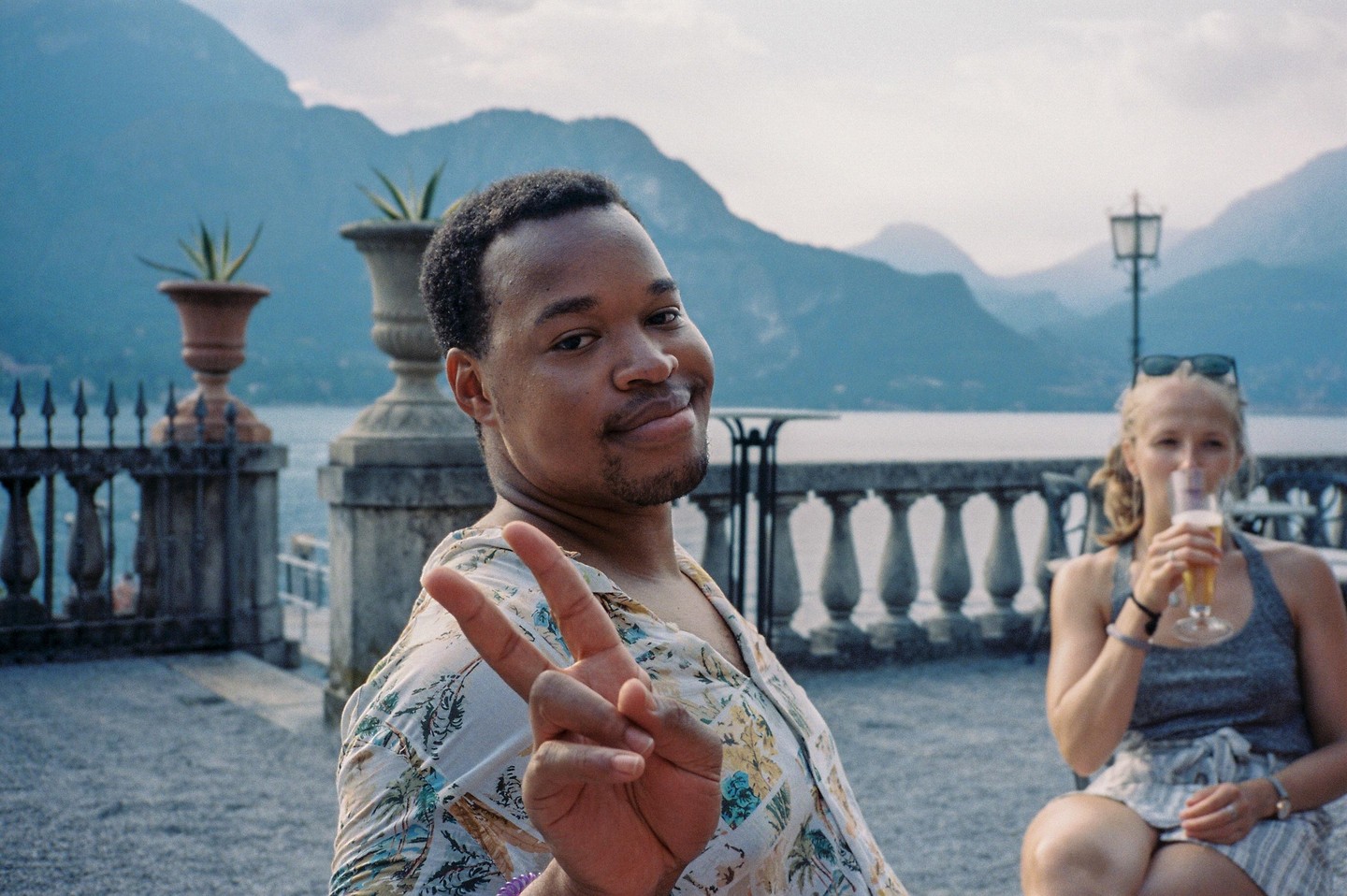 A man makes a peace sign while a woman drinks a beverage in the background, with a lake and mountains behind them.