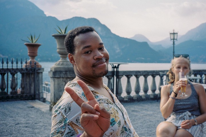 A man makes a peace sign while a woman drinks a beverage in the background, with a lake and mountains behind them.