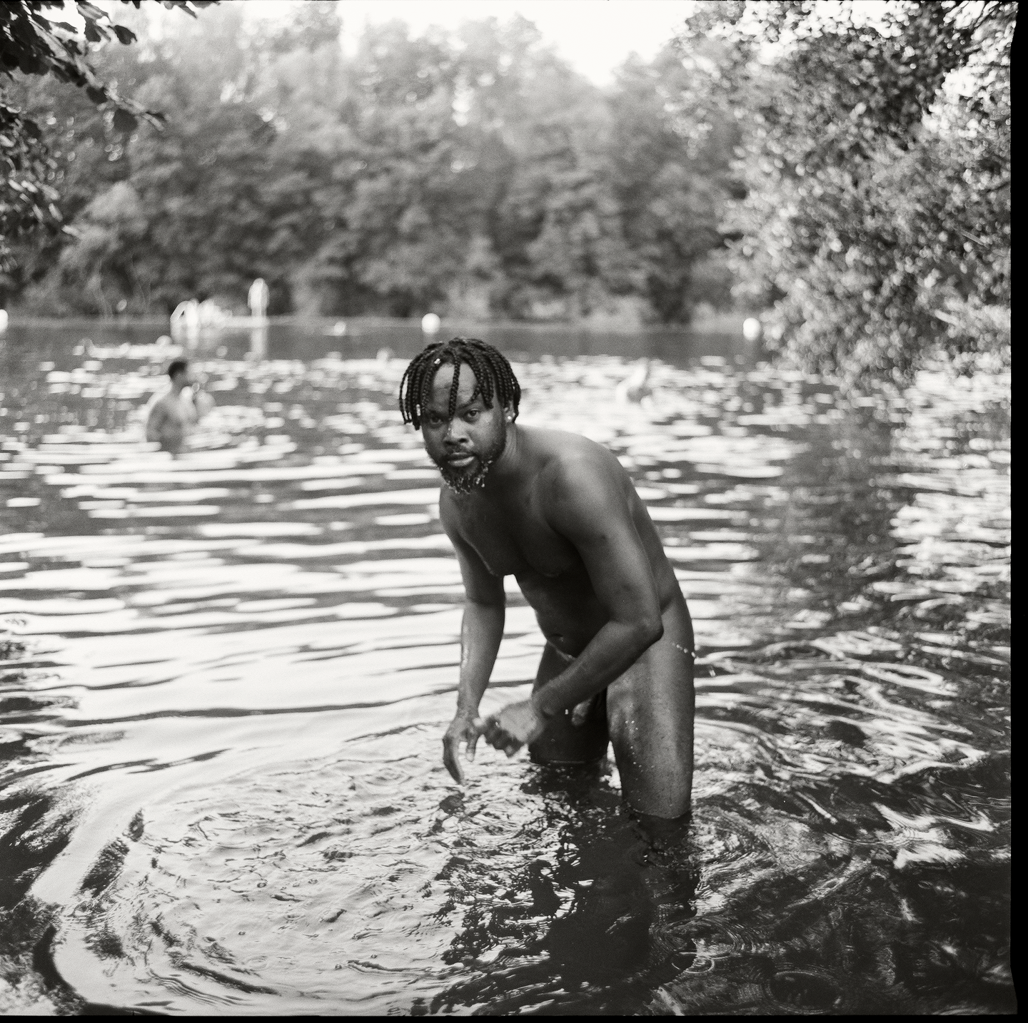 A man with braided hair stands in a lake with trees in the background.