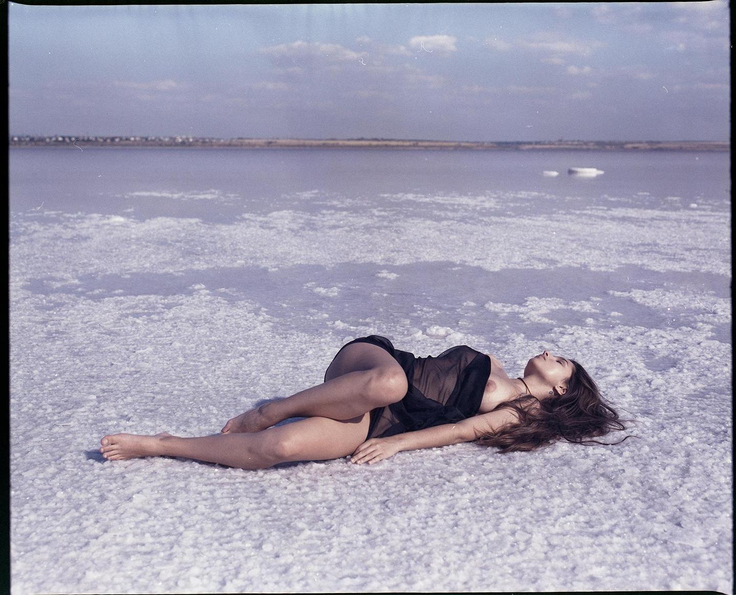A woman lies on a salt flat with a body of water and a distant shore in the background.