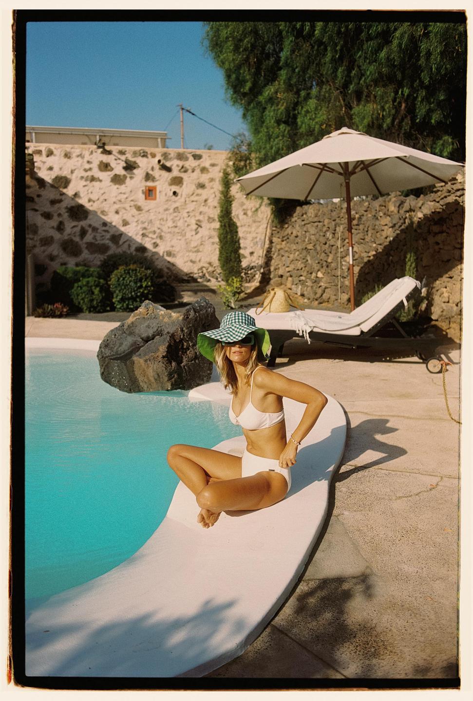 A woman in a white bikini and a green and white checkered hat sits by a swimming pool.