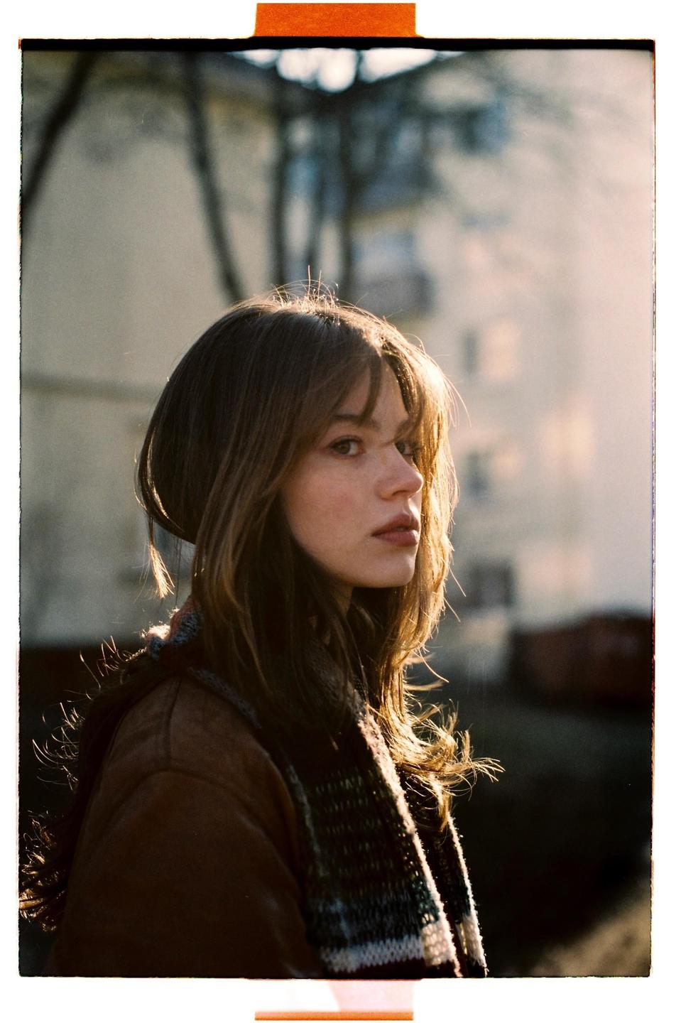 A woman with long hair and a scarf looks to the side with a building in the background.