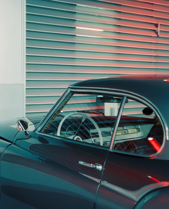 A vintage car with a white steering wheel is parked in front of a corrugated metal wall with red and blue lighting.