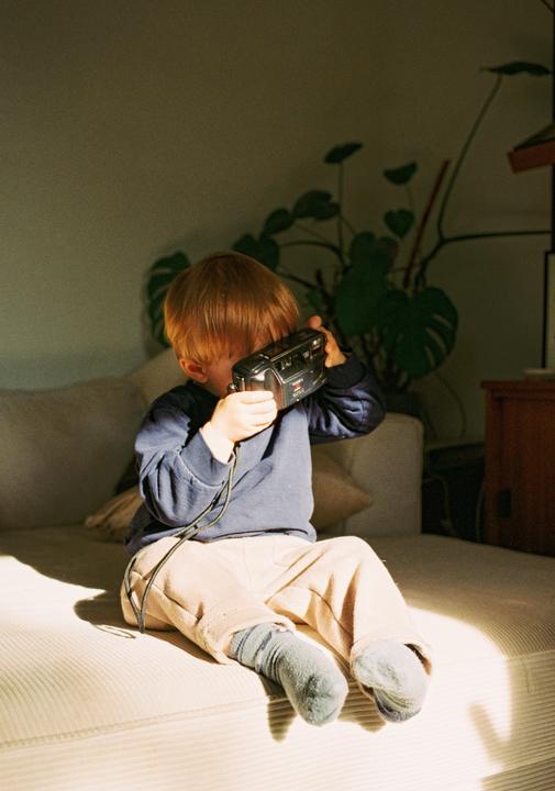 A young boy sits on a couch and holds a camera up to his face.