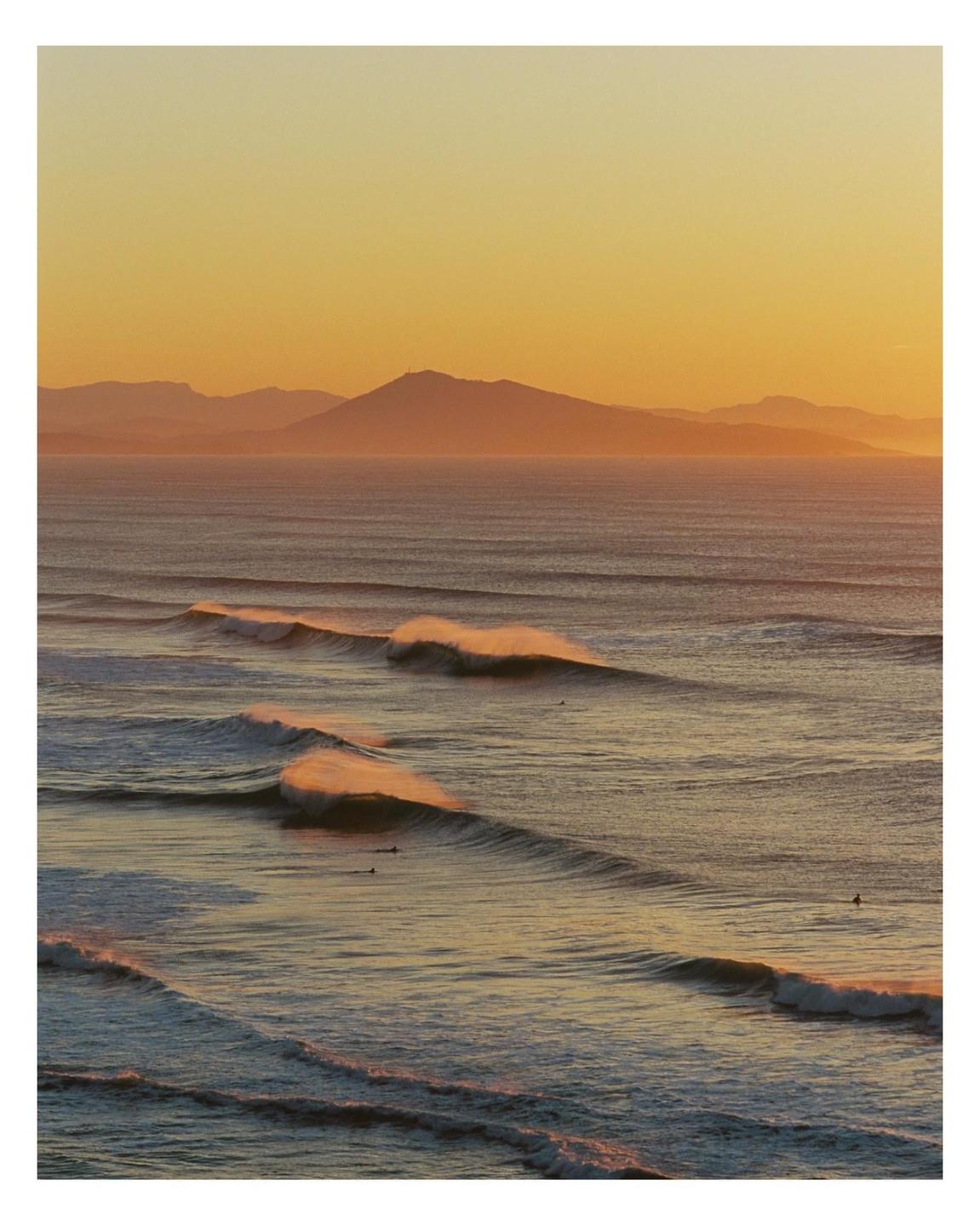 Waves roll towards the shore with mountains in the distance under an orange sky.