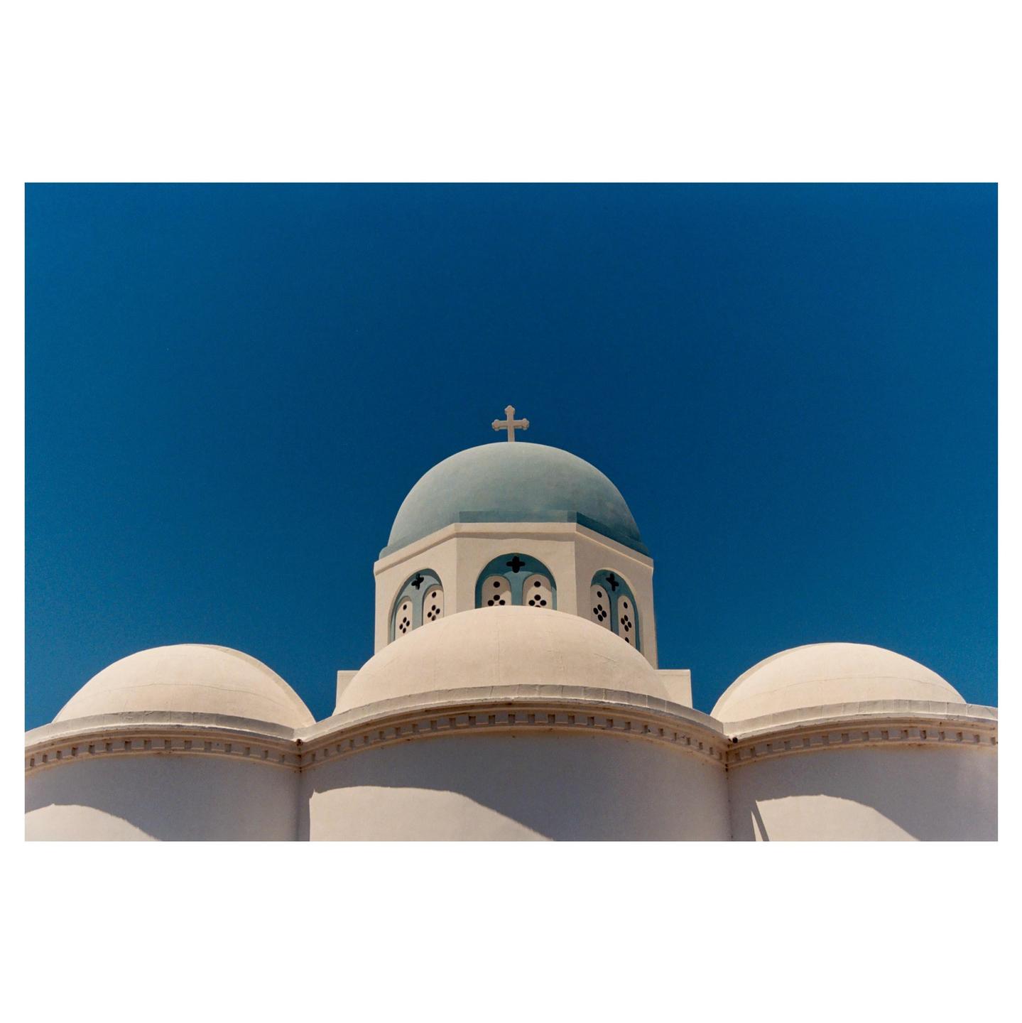 A white church with a blue dome and a cross on top stands against a clear blue sky.