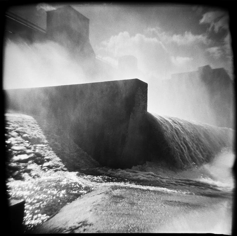 Water flows over a concrete dam, creating mist and spray.