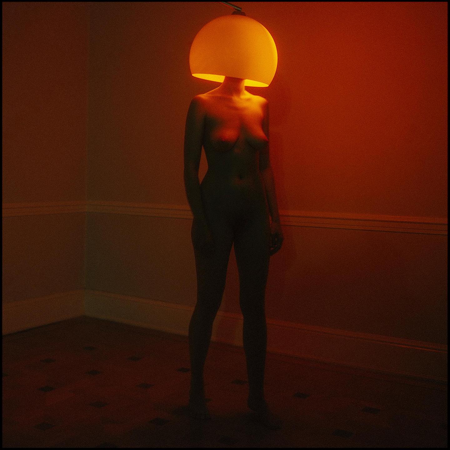 A nude woman stands in a corner with a lampshade covering her head, illuminated by a warm glow.