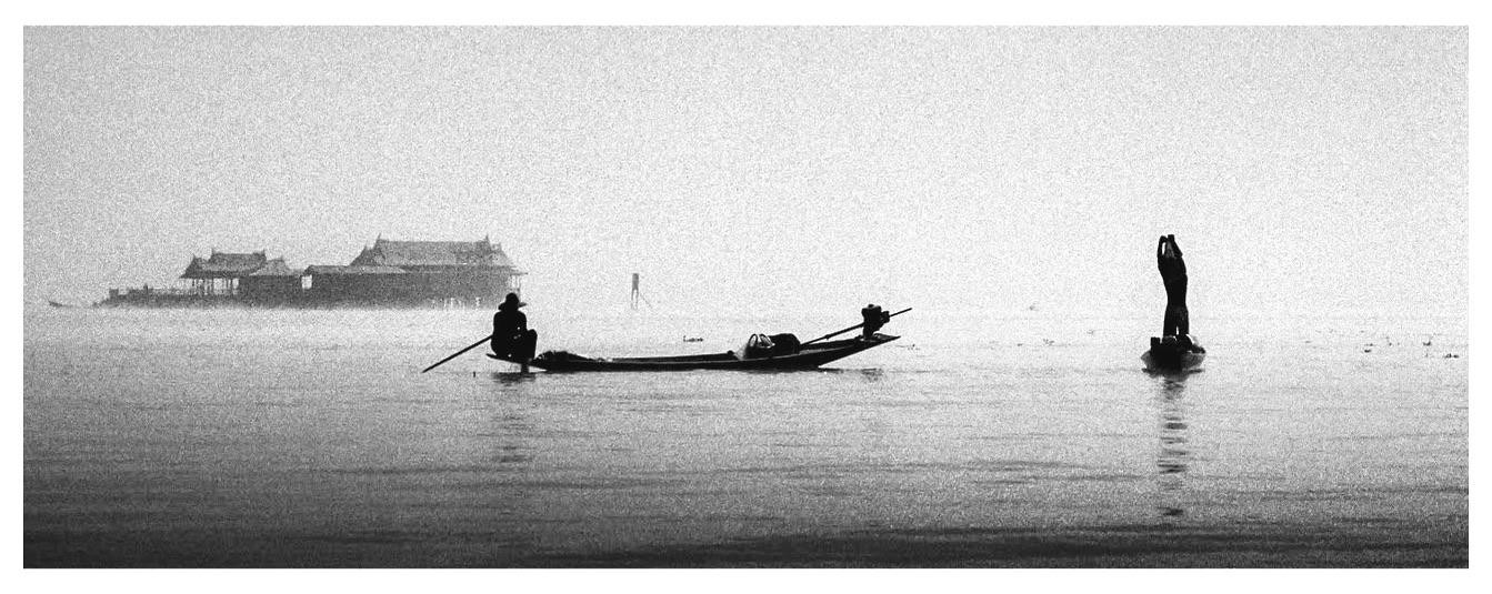 Two fishermen in boats are on a hazy lake with buildings in the background.