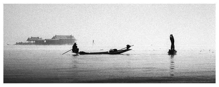 Two fishermen in boats are on a hazy lake with buildings in the background.