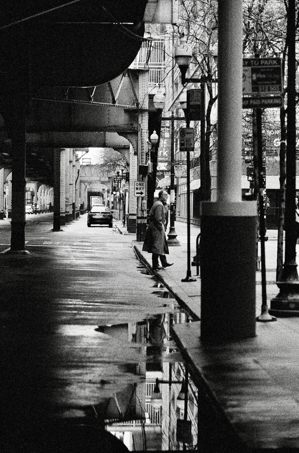 A man stands on a wet sidewalk next to a street with a car and an elevated train track overhead.