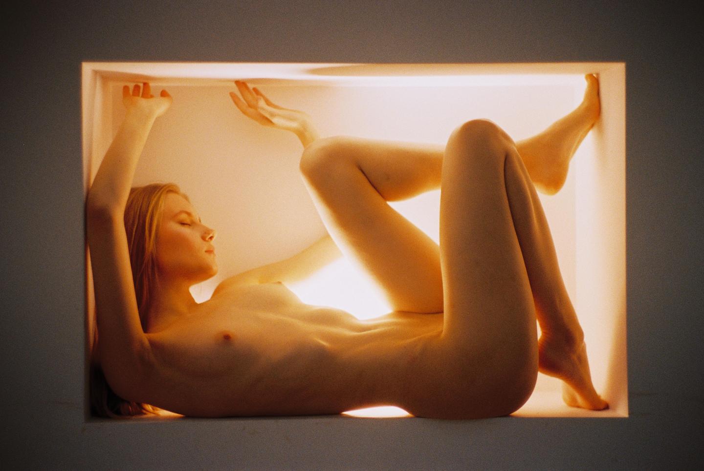 A nude woman is lying on her back inside a brightly lit rectangular alcove with her arms and legs bent.
