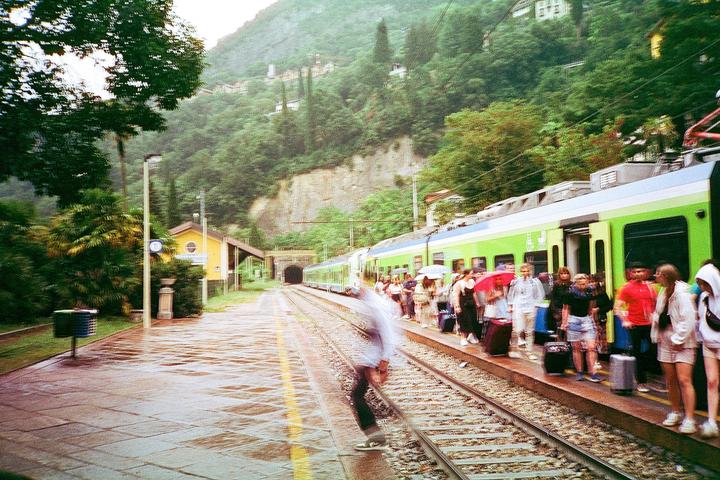 A train is stopped at a station platform with people waiting to board, next to a tunnel entrance and a lush green mountain.