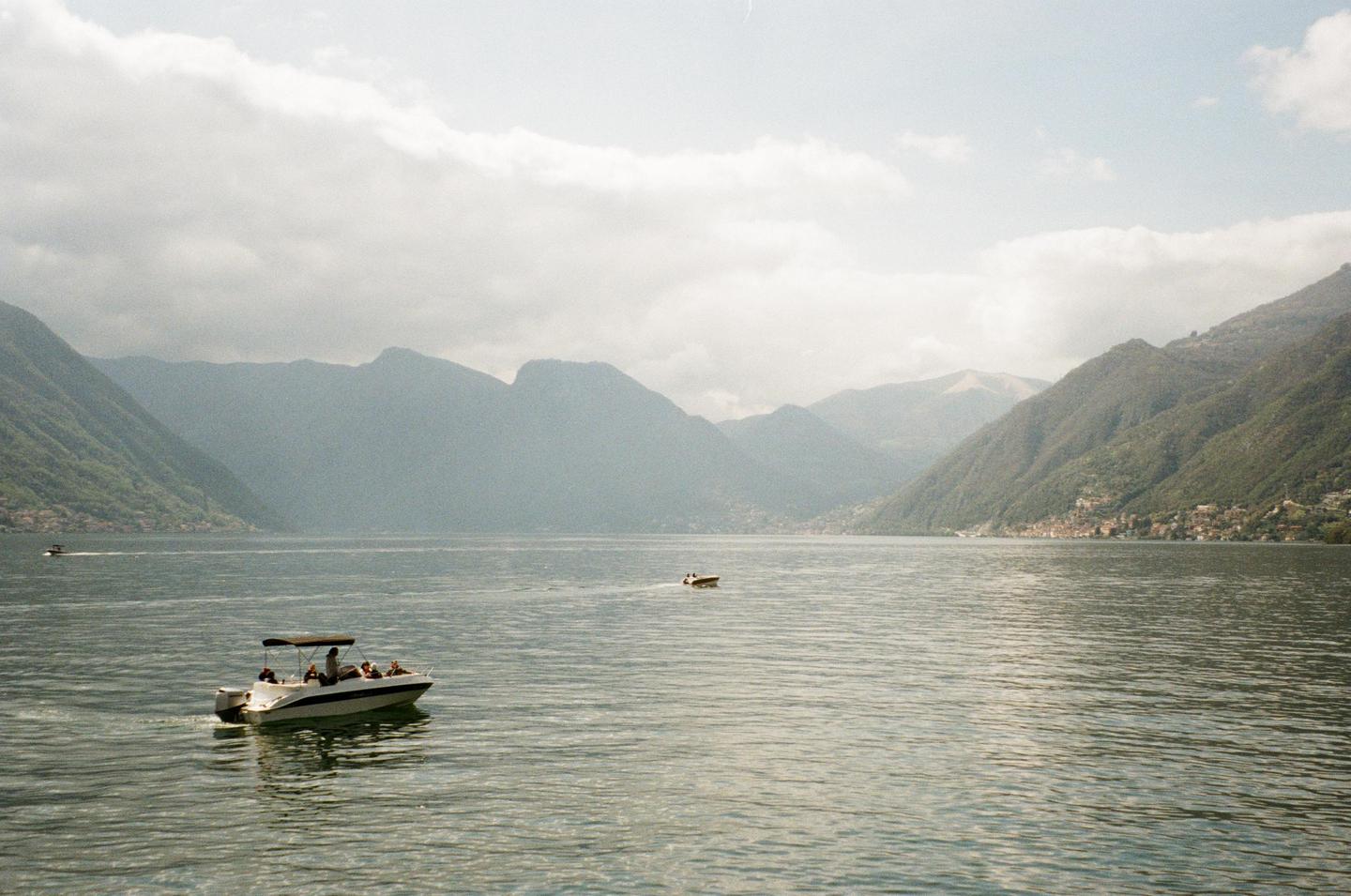 A motorboat with people on board cruises on a lake surrounded by mountains under a cloudy sky.