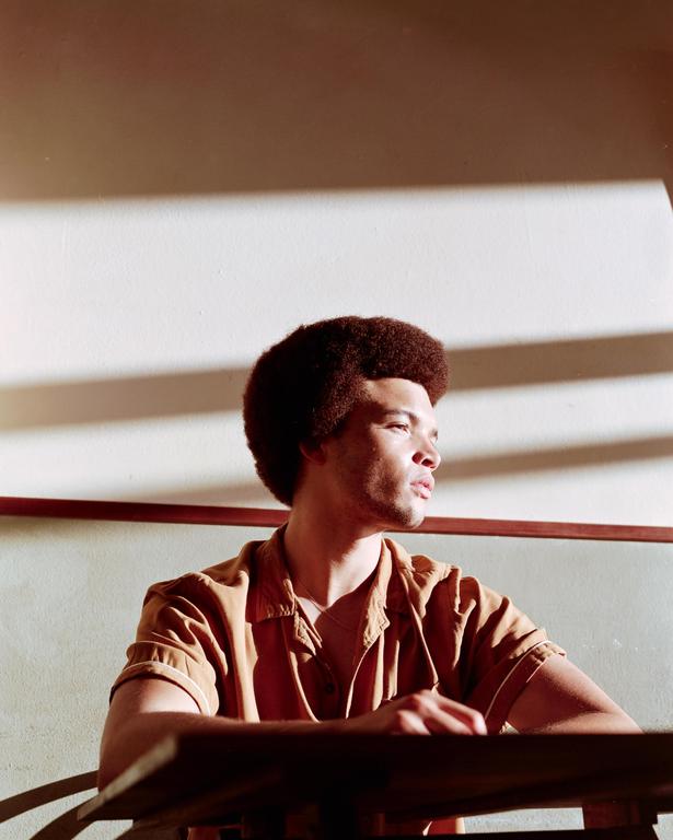 A man with an afro sits at a table with sunlight and shadows on the wall behind him.
