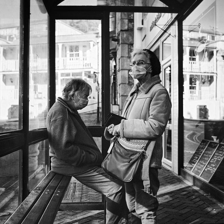 Two women wearing face masks are at a bus stop, one sitting on a bench and the other standing.