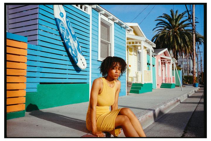 A woman in a yellow dress sits on a curb in front of colorful houses with a surfboard mounted on one of them.