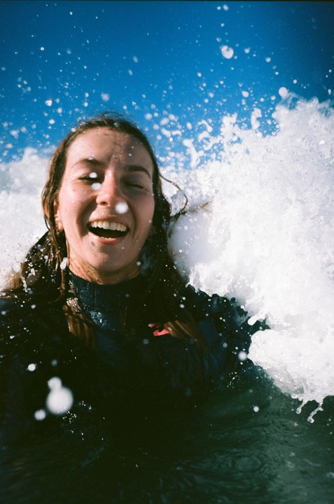 A woman with closed eyes and an open mouth is surrounded by splashing water under a blue sky.