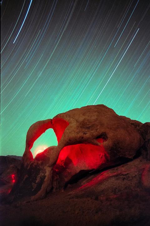 A rock formation with an arch is illuminated in red under a night sky with star trails.