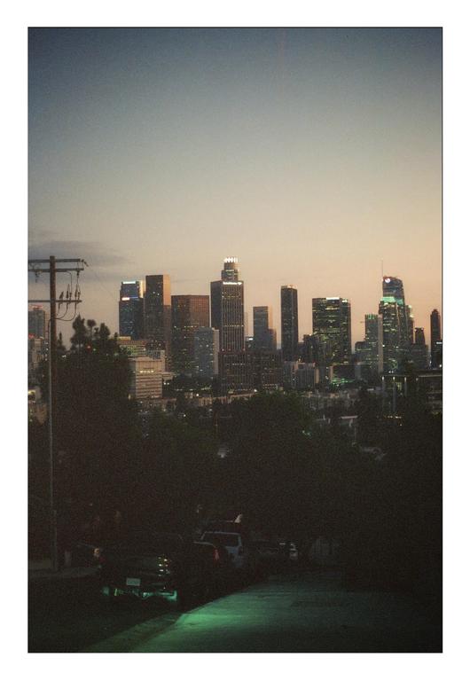A city skyline with illuminated buildings is visible above a dark street and trees at dusk.