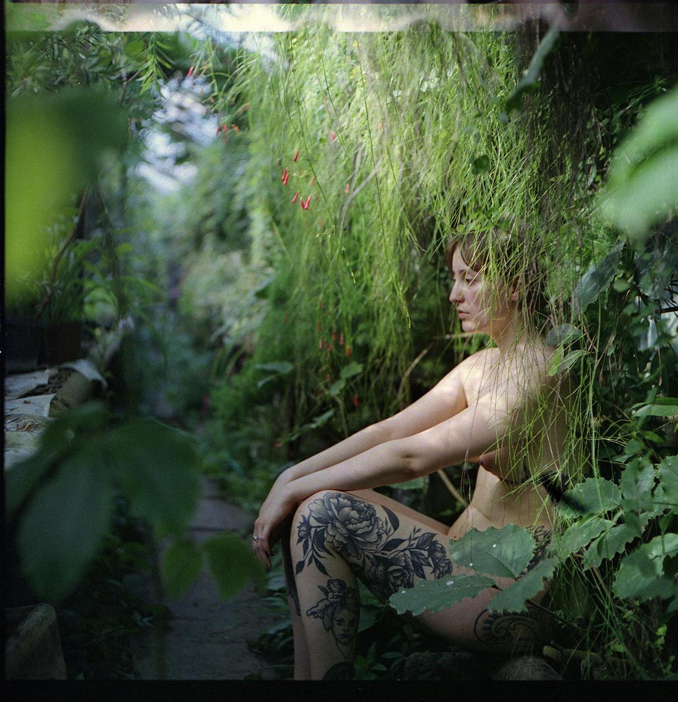 A woman with tattoos on her leg sits among lush green foliage.