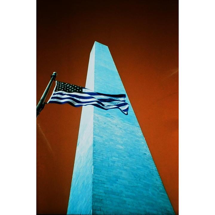 An American flag flies in front of the Washington Monument against an orange sky.