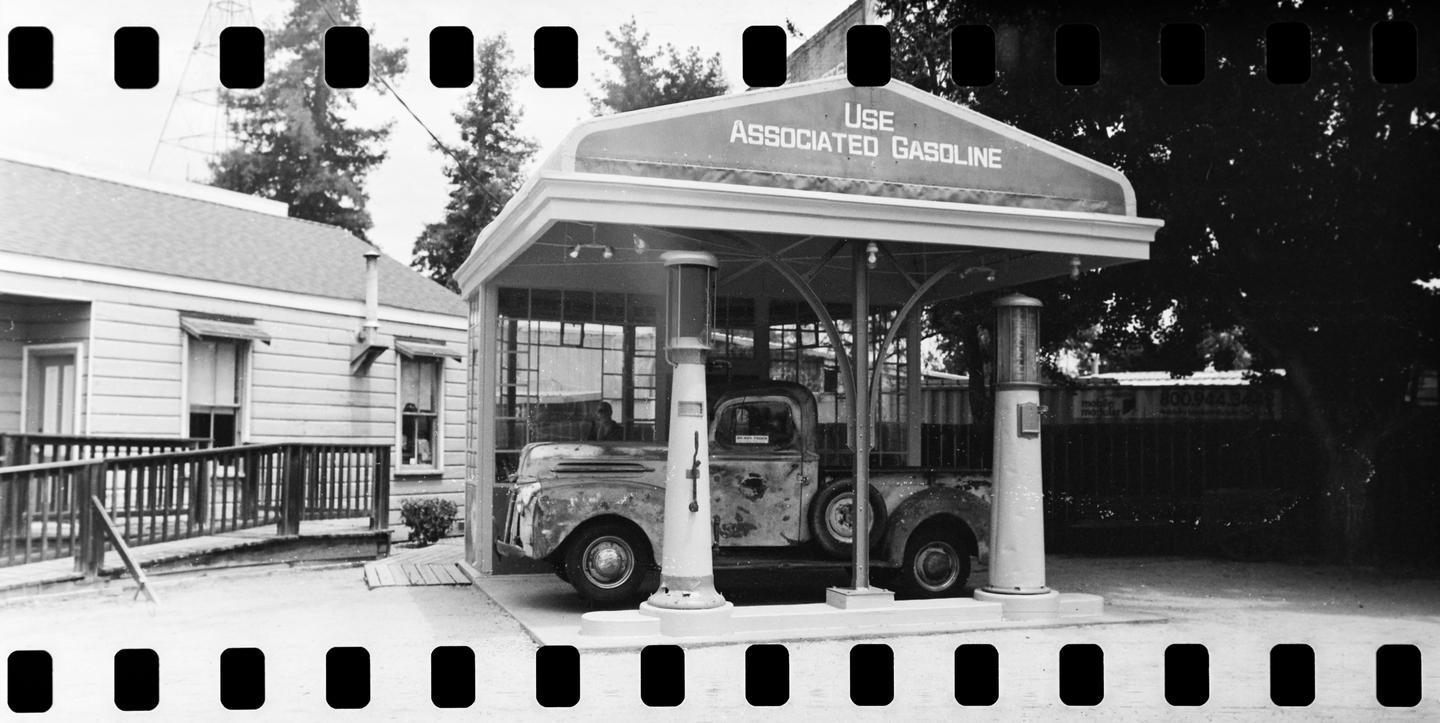A black and white photograph shows a vintage gas station with a truck parked underneath its canopy.