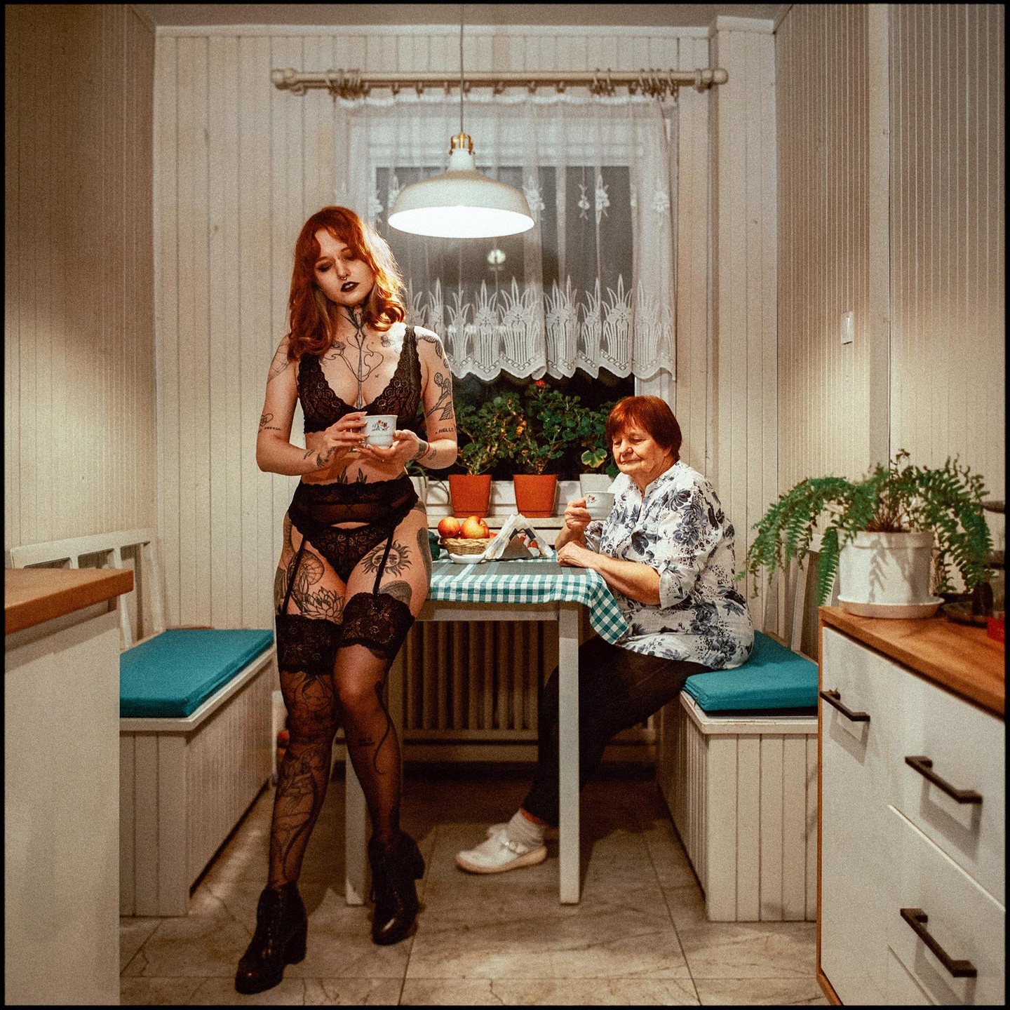 A woman in lingerie stands next to a table where an older woman is seated, both holding cups.