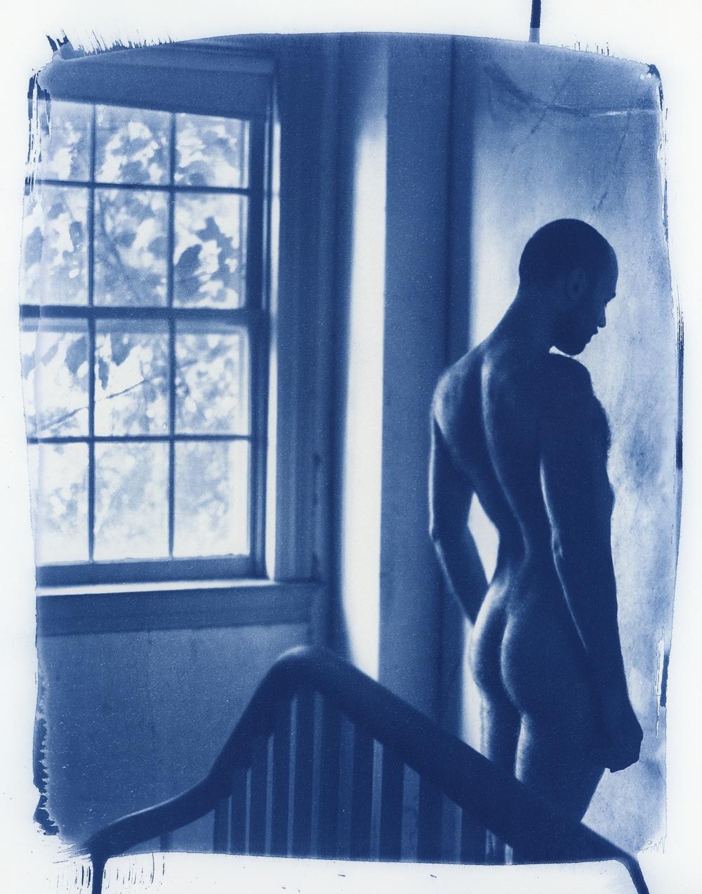 A nude man stands next to a window with a bed frame in the foreground.