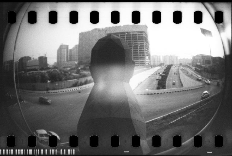 A fisheye view shows a statue overlooking a multi-lane road with cars and buildings in the background.