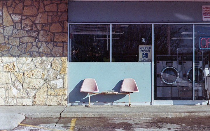 Two pink chairs sit outside a laundromat with washing machines visible through the window.