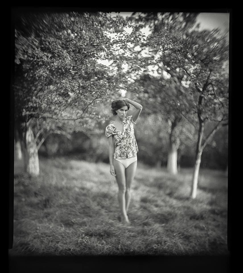 A woman stands barefoot in a field with trees in the background.