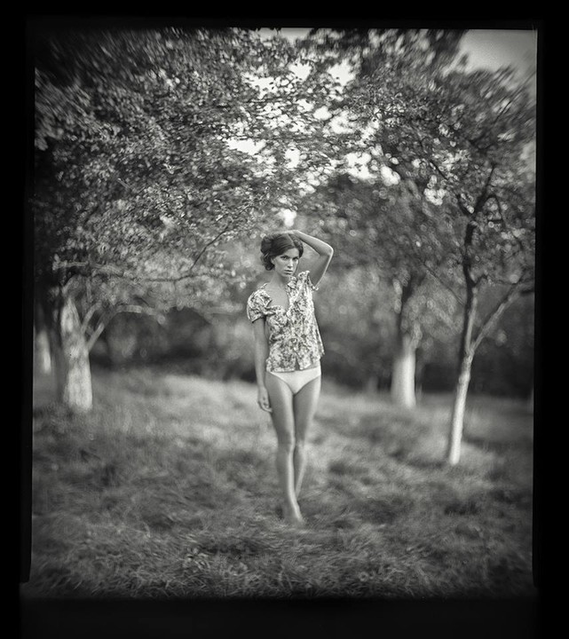 A woman stands barefoot in a field with trees in the background.