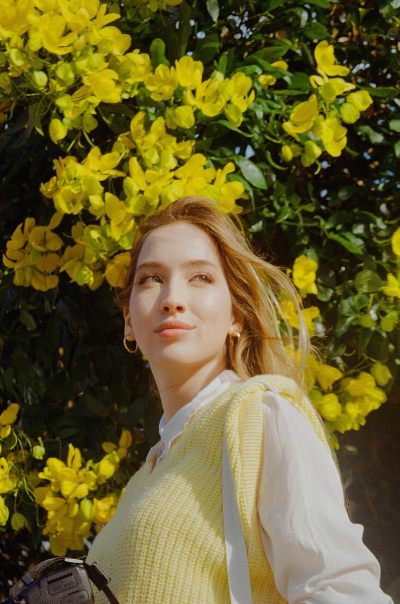 A woman with long hair and hoop earrings looks up, standing in front of a bush with yellow flowers.